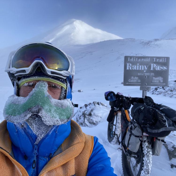 Peter Delamere poses in front of a sign in Rainy Pass in the Alaska Range during the 2024 Iditarod Trail Invitational race. On his face is a “nose-hat” invented by Fairbanks athlete Shalane Frost. Photo by Peter Delamere.